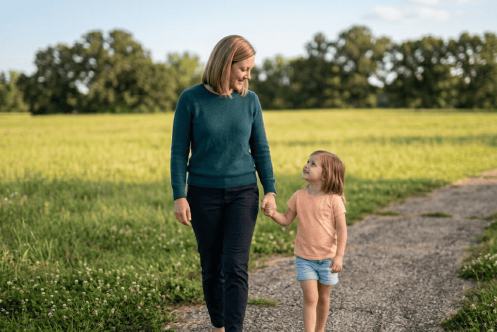 Mother and young daughter holding hands while walking on a gravel path through a sunlit field park.